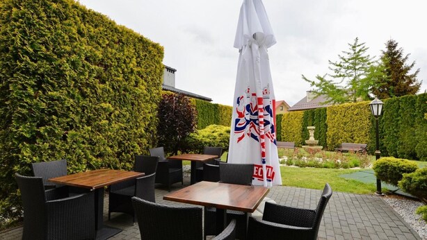 An outdoor seating area with tables and chairs, a white umbrella, and a hedge in the background.