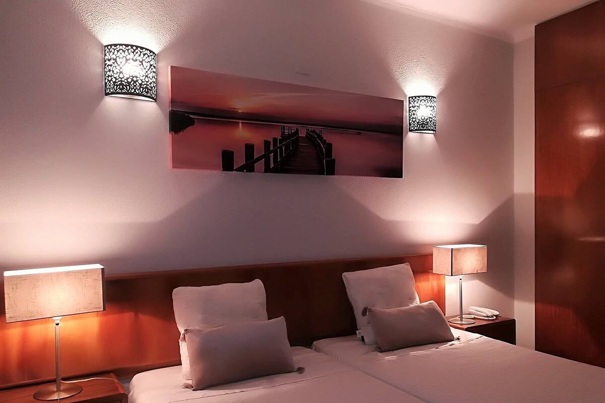 A hotel room with two beds, a large painting above the beds, and two wall-mounted lamps.