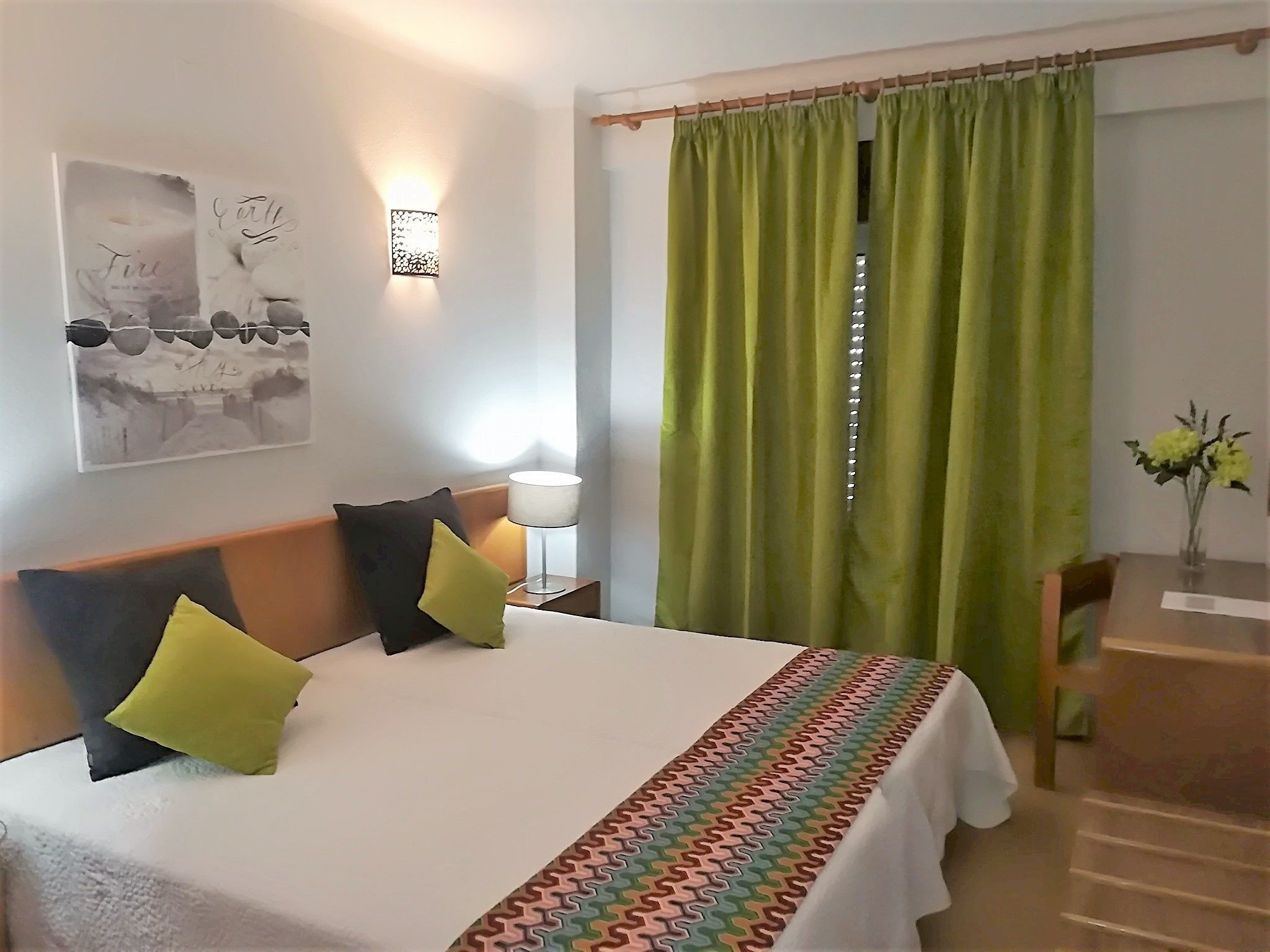 Hotellihuone, jossa on sänky, yöpöytä lampun kanssa, vihreä verho ja pieni pöytä, jonka päällä on maljakko täynnä kukkia.