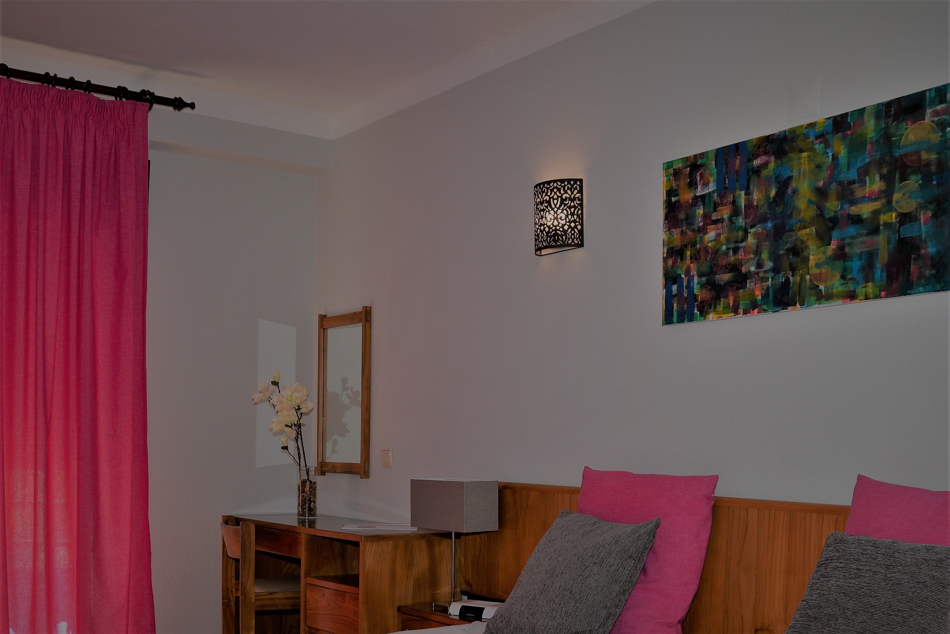 Un dormitorio con cabecera de madera, cama con almohadas rosadas y grises, un escritorio con lámpara, una cortina rosa y un cuadro abstracto colorido en la pared.