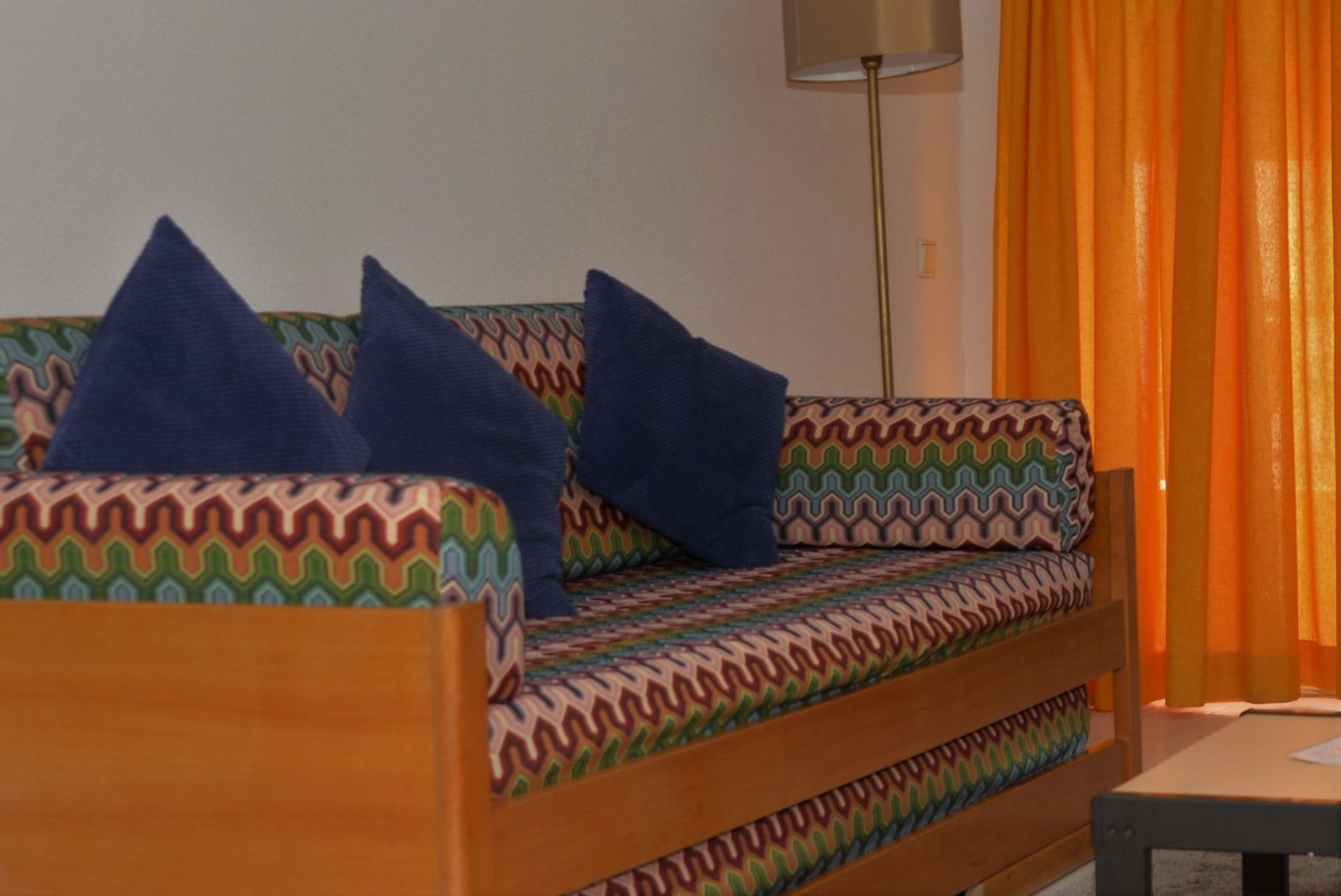 A sofa with a patterned fabric and two blue pillows, a floor lamp, and orange curtains.