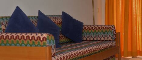 A sofa with a patterned fabric and two blue pillows, a floor lamp, and orange curtains.