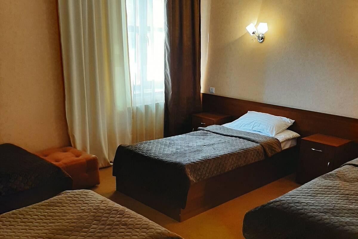 A hotel room with three beds arranged in a row, a nightstand, a wall-mounted light, and a window with curtains.