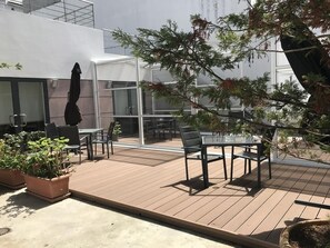 An outdoor patio area with a wooden deck, a glass table, and chairs. There are potted plants and a large tree providing shade.