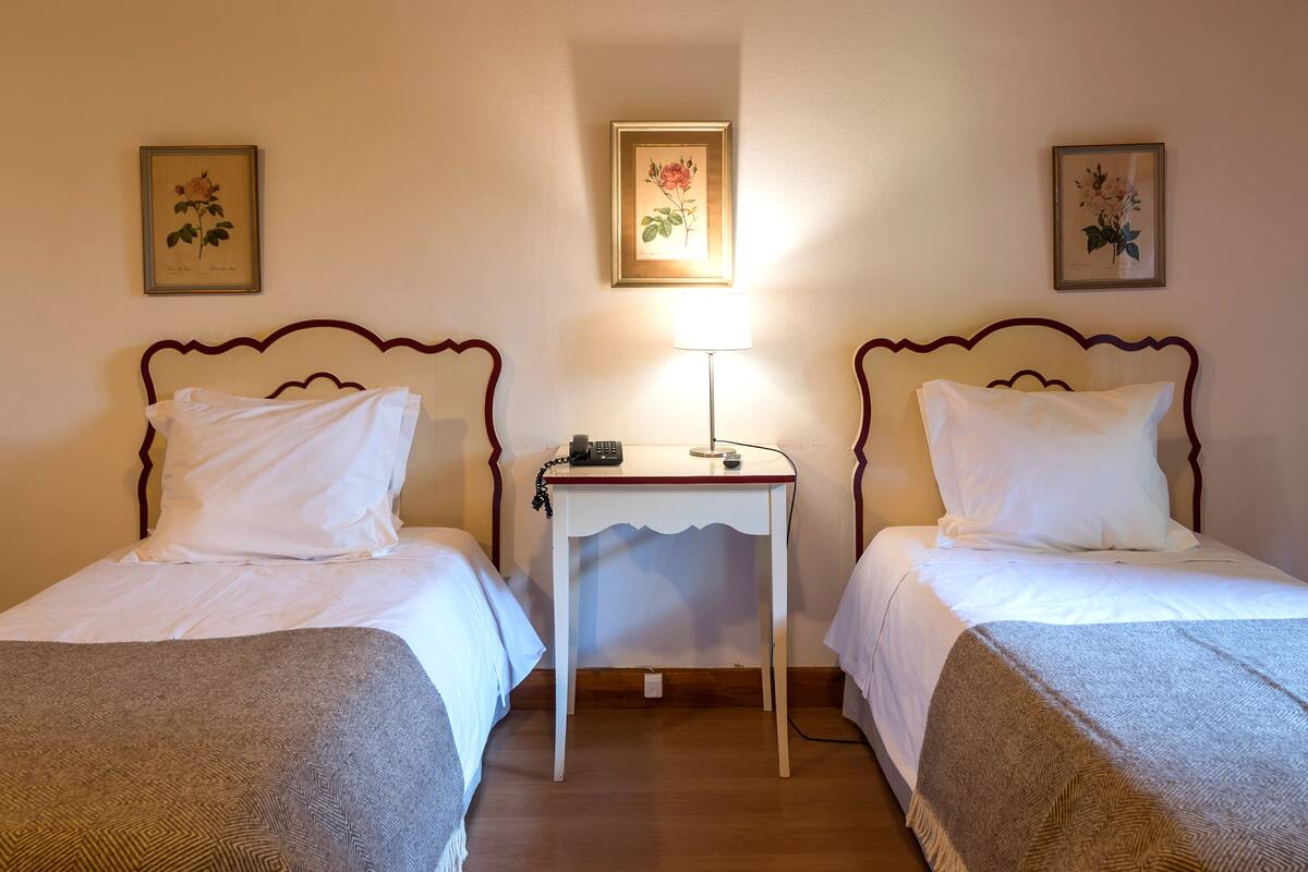 A hotel room with two single beds, a small bedside table with a lamp, and framed botanical prints on the walls.