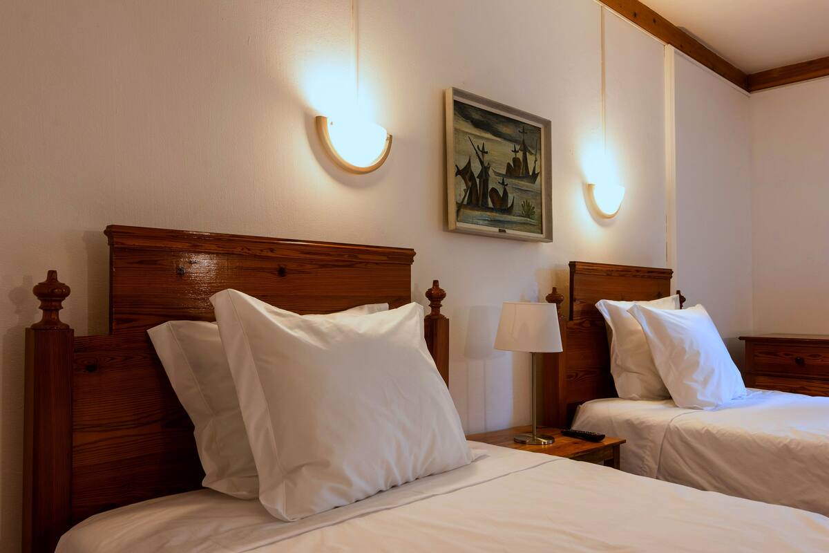 Two wooden beds with white linens and headboards, a nightstand with a lamp, and a framed painting on the wall.