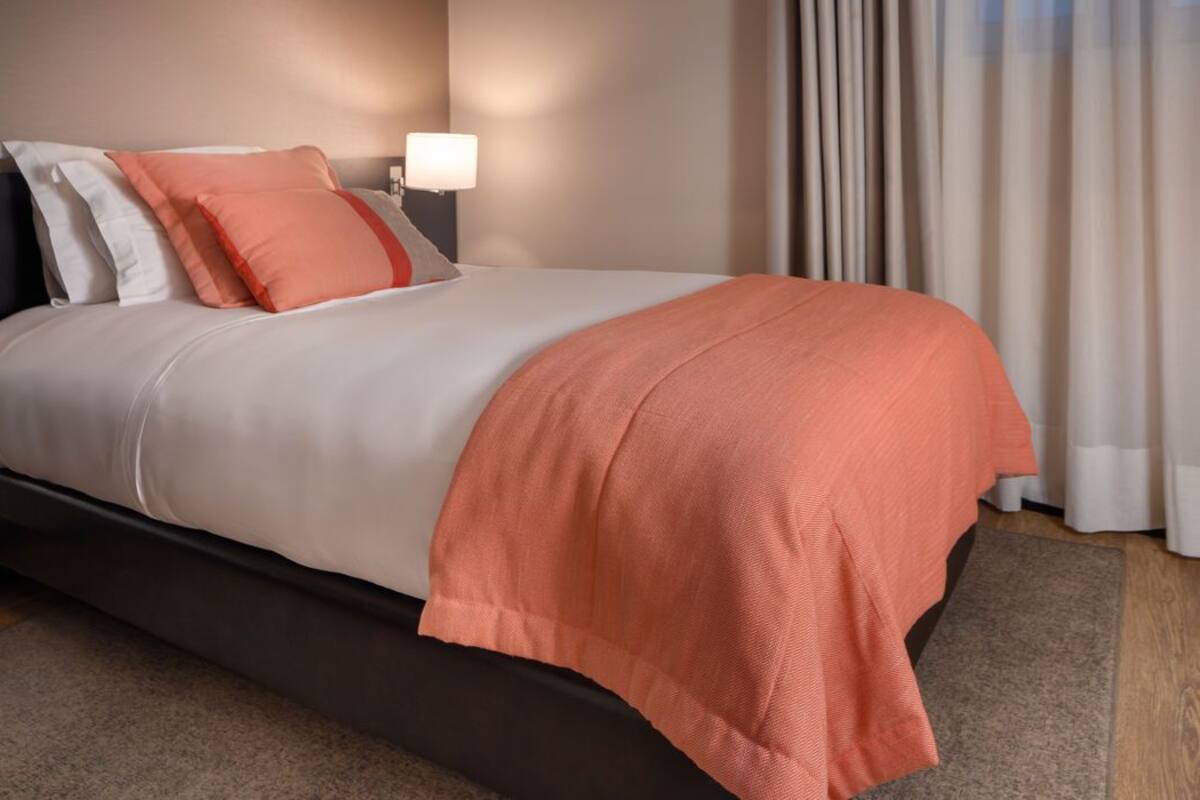Ein ordentlich bezogenes Bett mit weiß-orangefarbener Bettdecke, einem dunklen Kopfteil und einer Nachttischlampe.