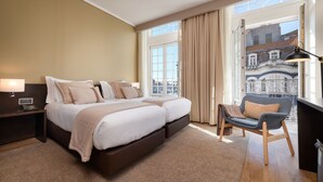 Een hotelkamer met een tweepersoonsbed, een bureau met lamp, een stoel en een groot raam met uitzicht op gebouwen.