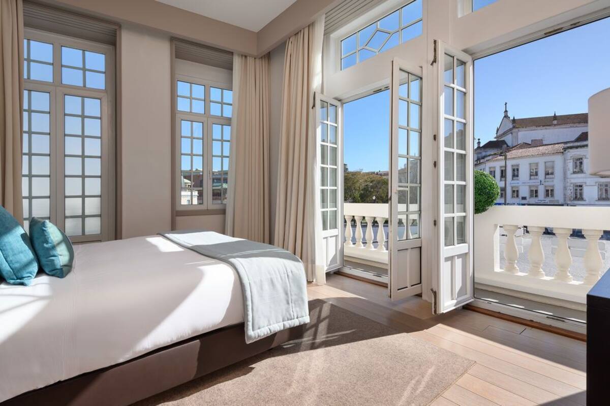 Ein Schlafzimmer mit einem großen Bett, einem Balkon mit Blick auf Gebäude und einem Fenster mit Vorhängen.