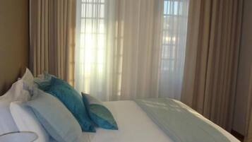 Una cama bien tendida con ropa de cama blanca y azul, un cabecero oscuro y una ventana con cortinas traslúcidas que dejan entrar luz natural.