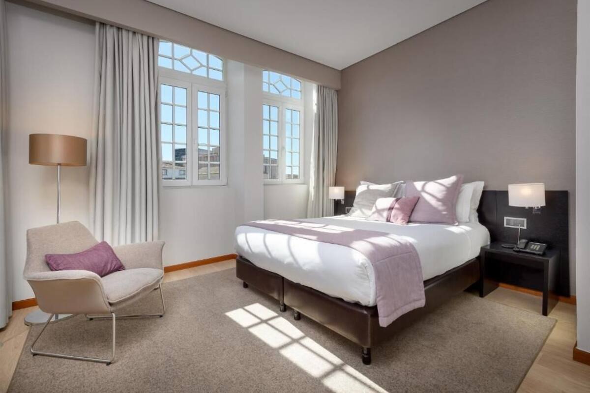 Ein modernes Hotelzimmer mit einem großen Bett, einem Sessel, zwei Nachttischen und Blick auf die Stadt durch große Fenster.