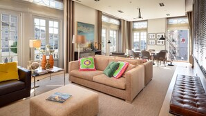 Een ruime woonkamer met een beige bank, een bruine leren zitbank en een salontafel. Grote ramen met gordijnen, een eethoek op de achtergrond, en een balkon met een hekwerk.
