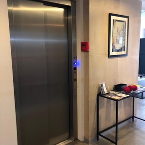 Um elevador com painel de controle, um botão de emergência vermelho, um quadro na parede e uma mesa com objetos no chão.