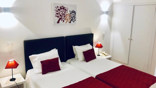 A hotel room with two beds, white bedding, red throw pillows, a red lamp on each side, a wall-mounted artwork, and a wardrobe. - Botânico (Coimbra)
