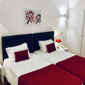 Quarto de hotel com duas camas, roupa de cama branca, almofadas decorativas vermelhas, mesa de cabeceira com abajur e uma obra de arte na parede.