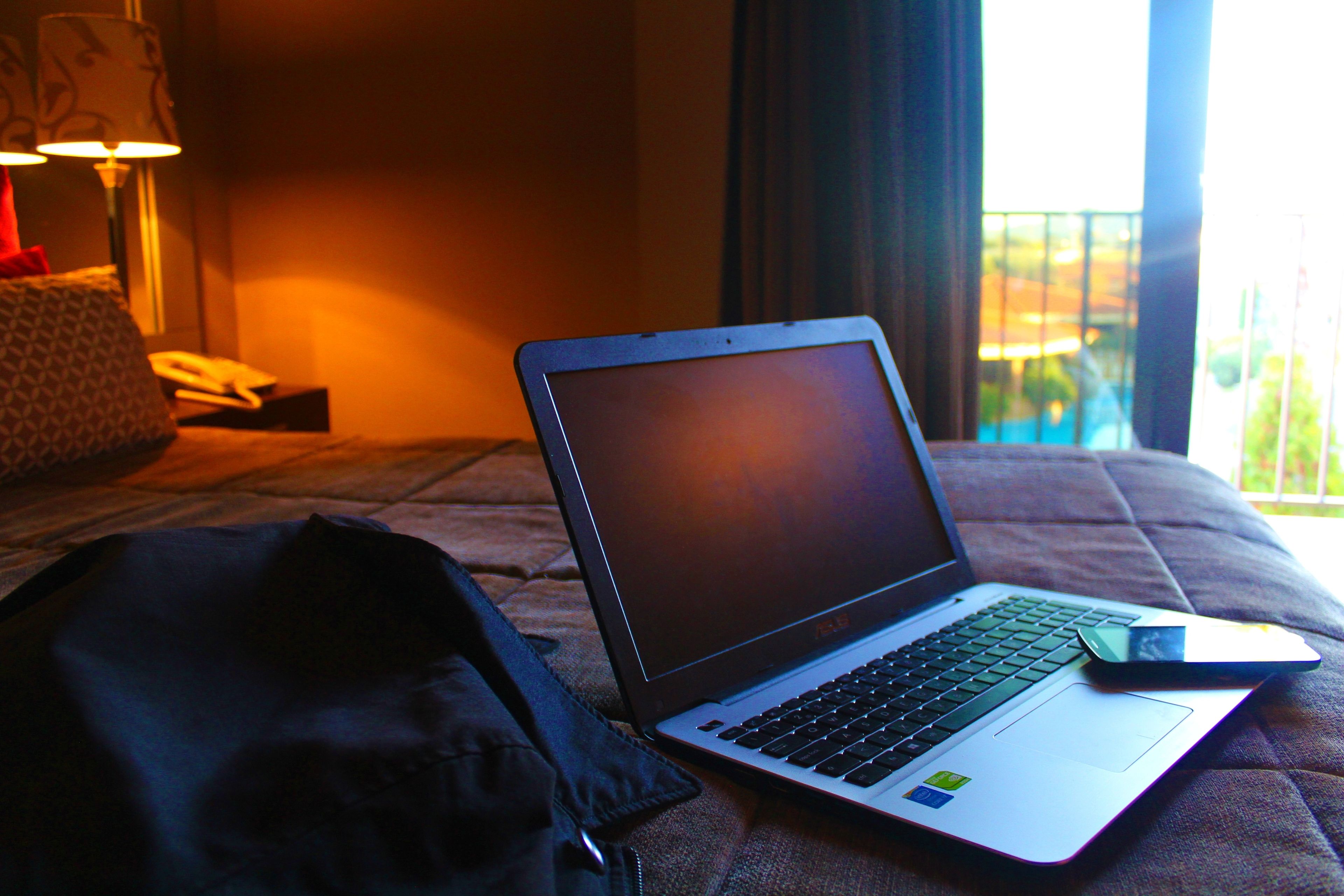Una laptop sobre una cama, con una manta, un almohadón y una lámpara al fondo.