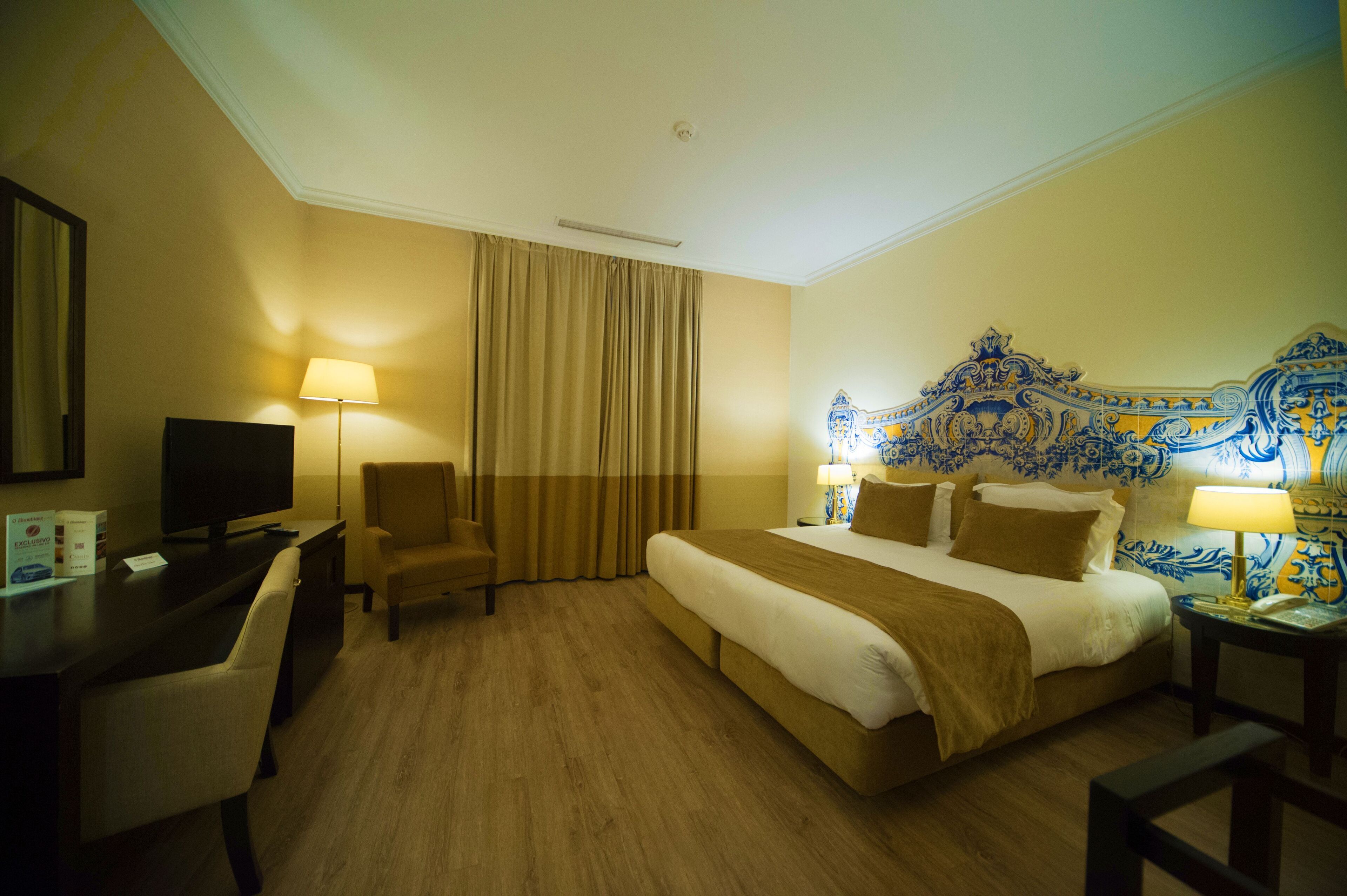 A hotel room with a large bed, a desk with a television, a chair, and a decorative headboard.