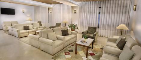 A spacious living room with multiple sofas and armchairs, a flat-screen TV mounted on the wall, and a large window with sheer curtains.