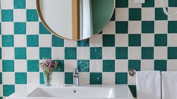 Un baño con salpicadero de azulejos a cuadros, un espejo redondo, un lavabo blanco, un florero con flores y un perchero.