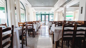 Een ruime eetzaal met hoge plafonds, witte betegelde vloeren en houten tafels en stoelen.