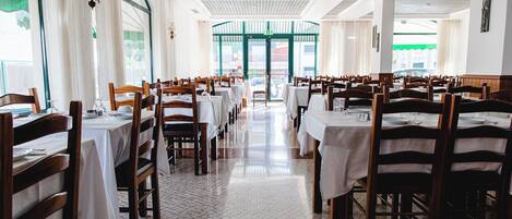 A spacious restaurant with high ceilings, white tiled floors, and wooden tables and chairs.