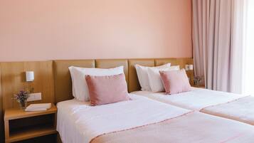 Habitación de hotel con dos camas, cada una con ropa blanca y almohadas rosas, cabeceros de madera, una mesita de noche con un florero y un ventanal con cortinas.