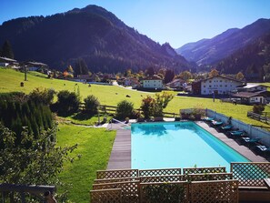 Un espace piscine doté d’une terrasse en bois et de chaises longues, entouré d’une clôture et de verdure, avec vue sur un village et des montagnes en arrière-plan.
