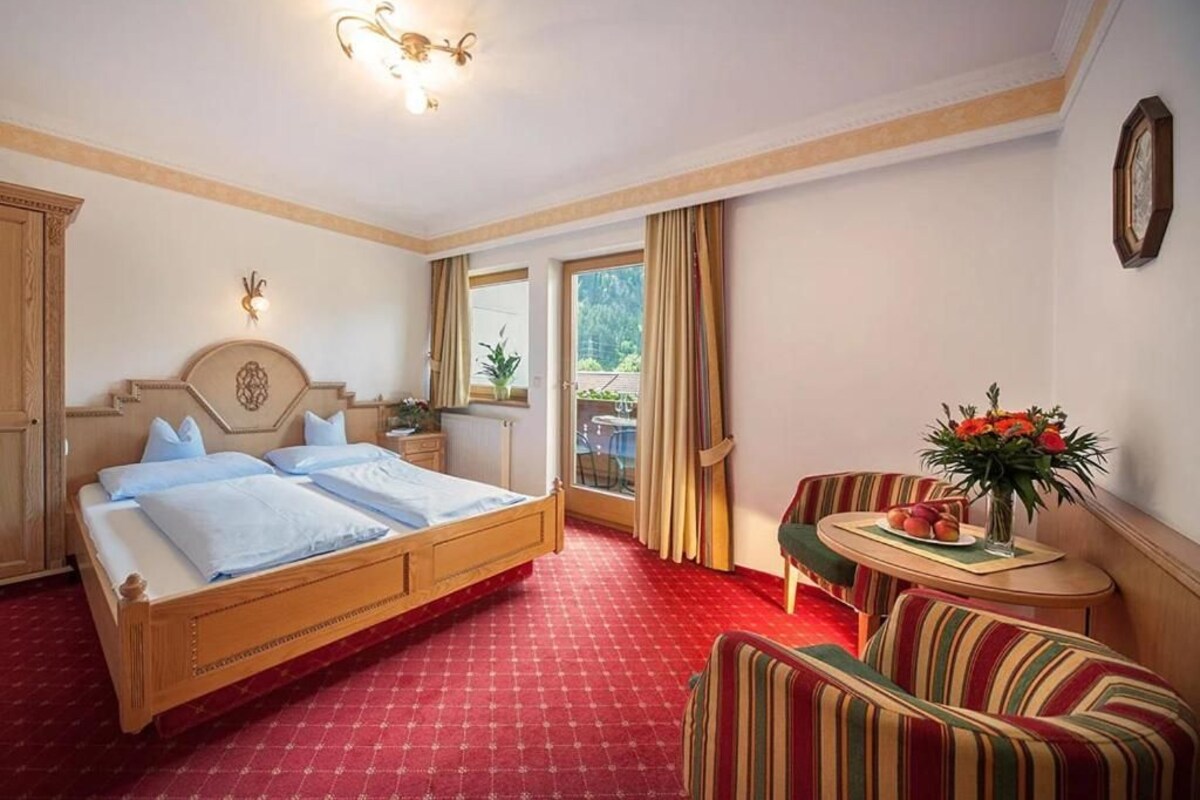A hotel room with a bed, a small table with flowers, a chair, and a view of the outdoors through a window.