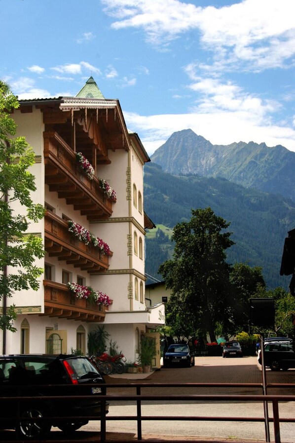 Un hôtel doté d’un balcon orné de fleurs, avec une chaîne de montagnes en arrière-plan et des voitures garées au premier plan.