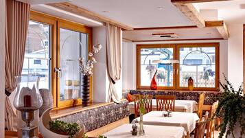 A dining area with a high wooden ceiling, large windows, and a view of the outside. There are two tables set for dining, a bench, and a decorative vase with flowers.