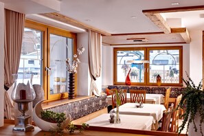 Un comedor con techo de madera alto, ventanas grandes y vistas al exterior. Hay dos mesas preparadas para comer, un banco y un jarrón decorativo con flores.