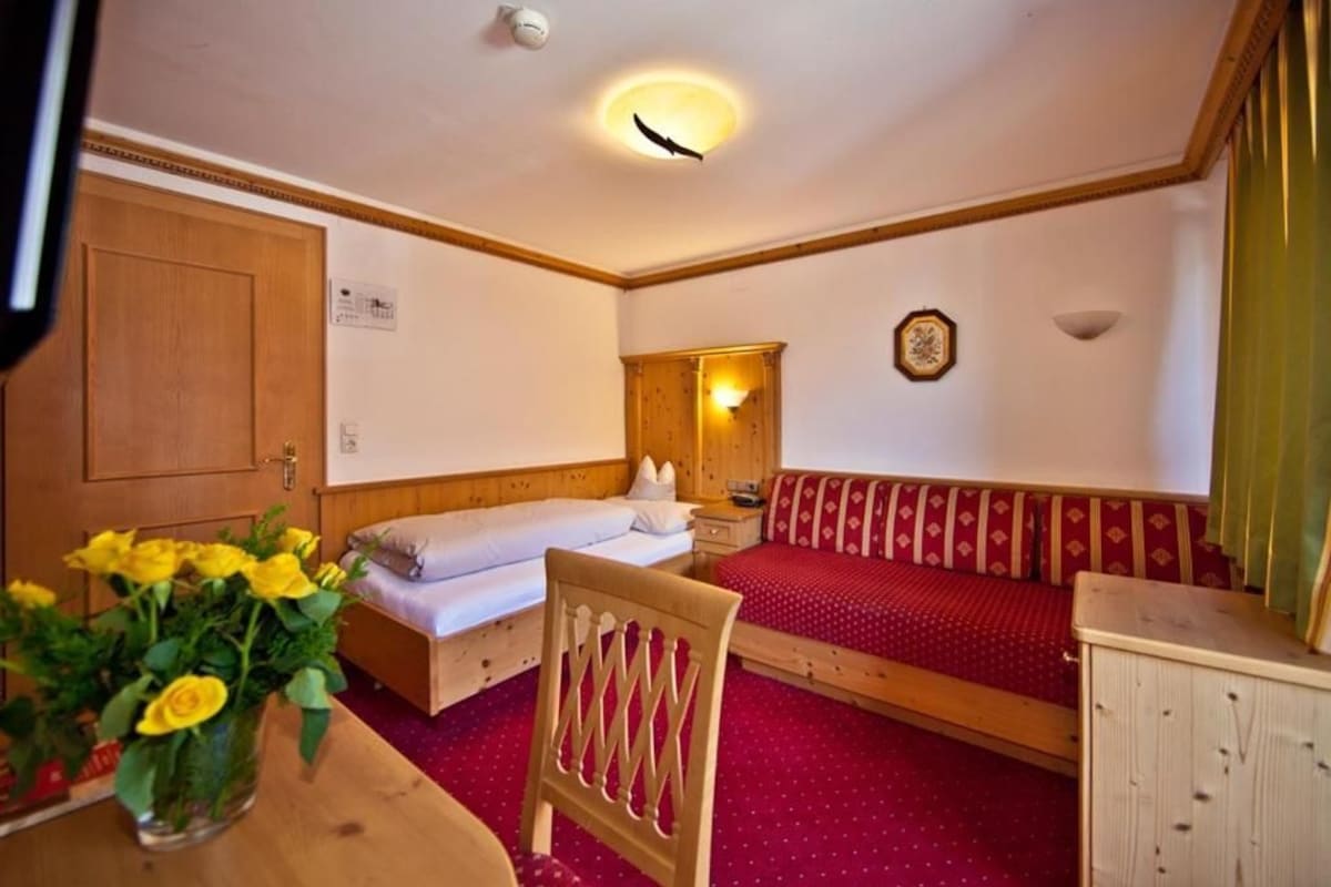 A room with two beds, a wooden chair, a table with a vase of flowers, and a clock on the wall.