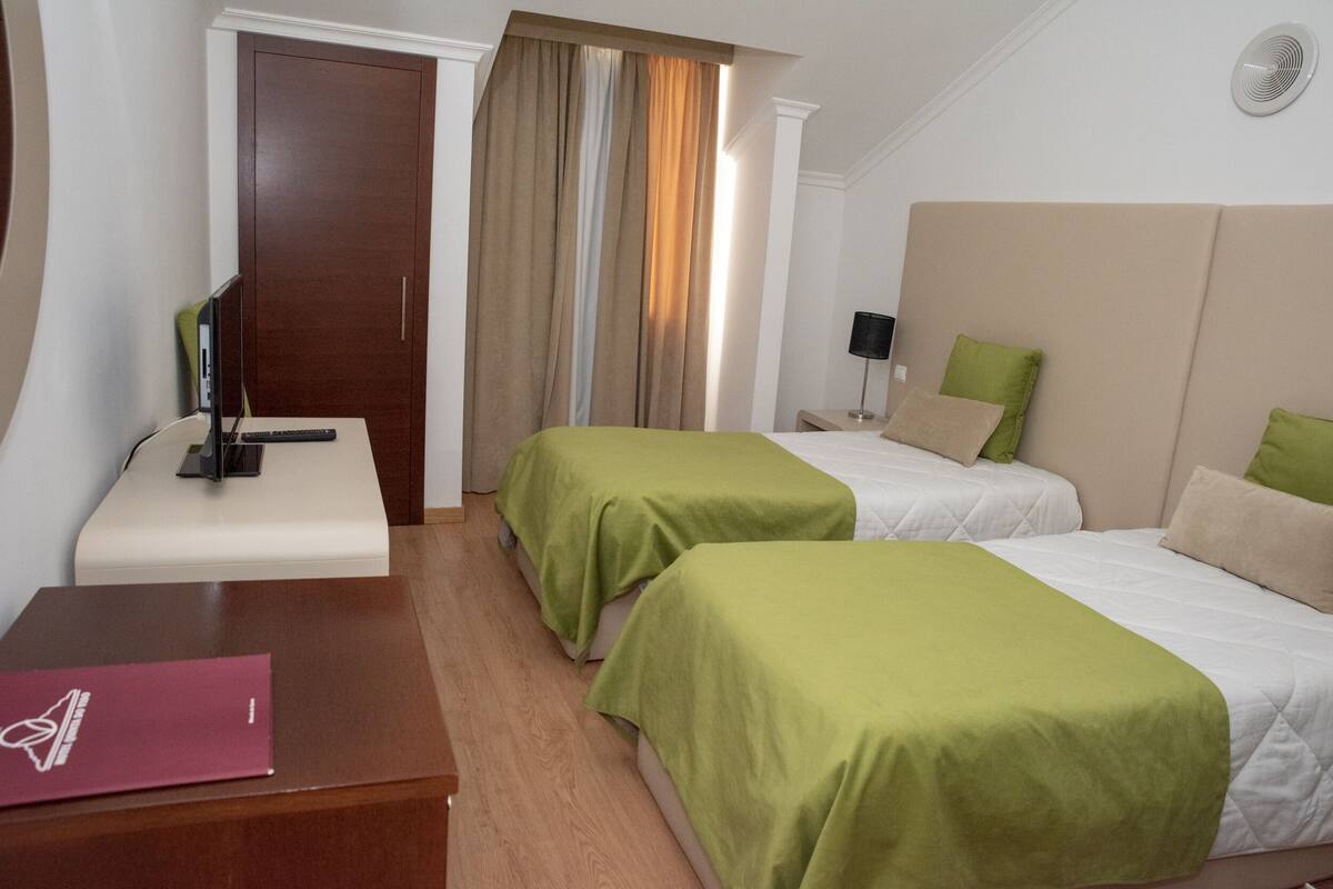 Ein Hotelzimmer mit zwei Einzelbetten, einem Schreibtisch mit Stuhl, einem Kleiderschrank und einem Fenster mit Vorhängen.