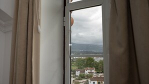 Een raam met gordijnen die gedeeltelijk zijn opengetrokken, met zicht op een woonwijk met huizen en een waterpartij in de verte.