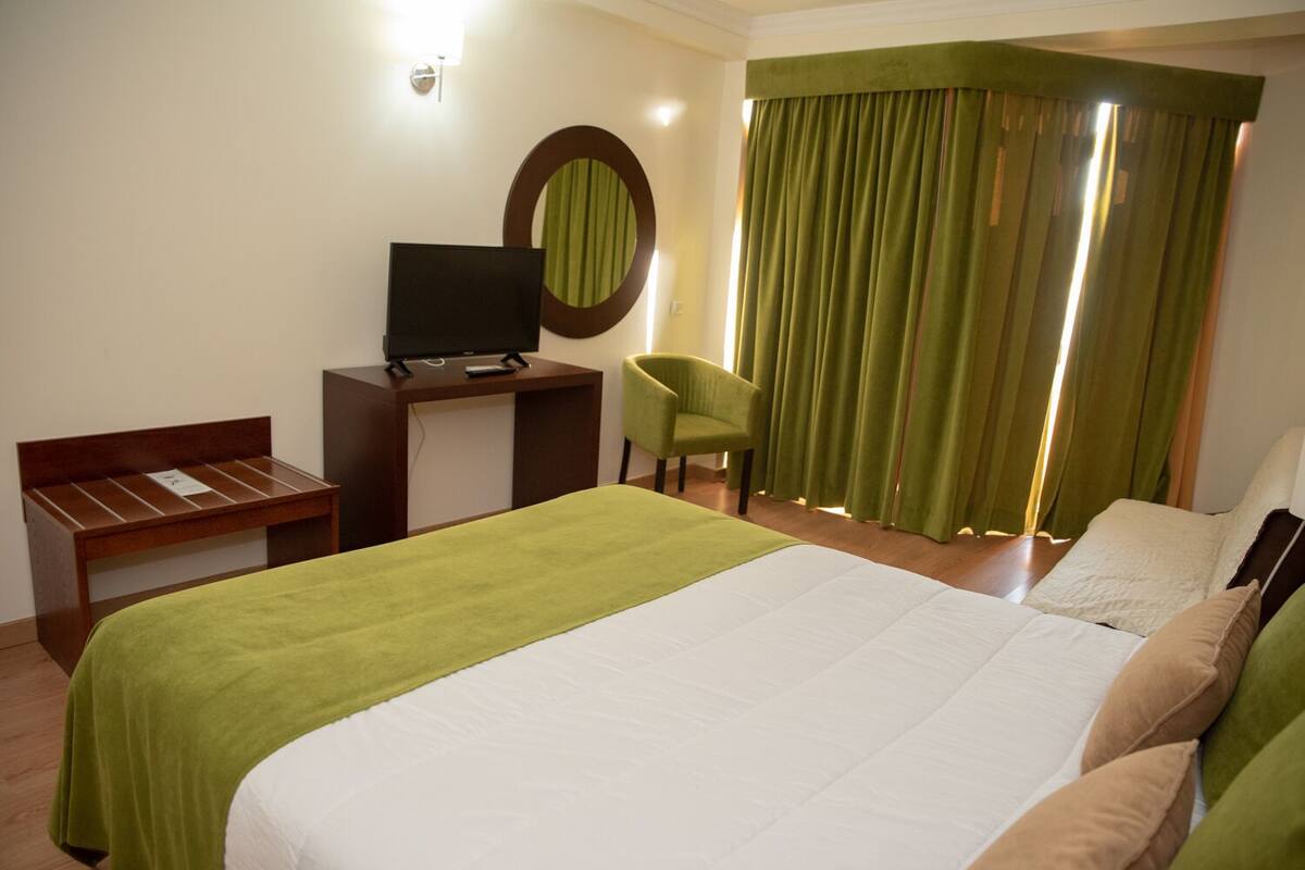 Ein Hotelzimmer mit einem großen Bett, einem Schreibtisch mit Fernseher, einem Sessel und einem Fenster mit grünen Vorhängen.