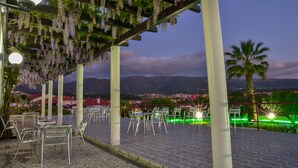 Een terras buiten met tafels en stoelen, een pergola met bloemen die eraan hangen, en een uitzicht op een stad en bergen op de achtergrond.