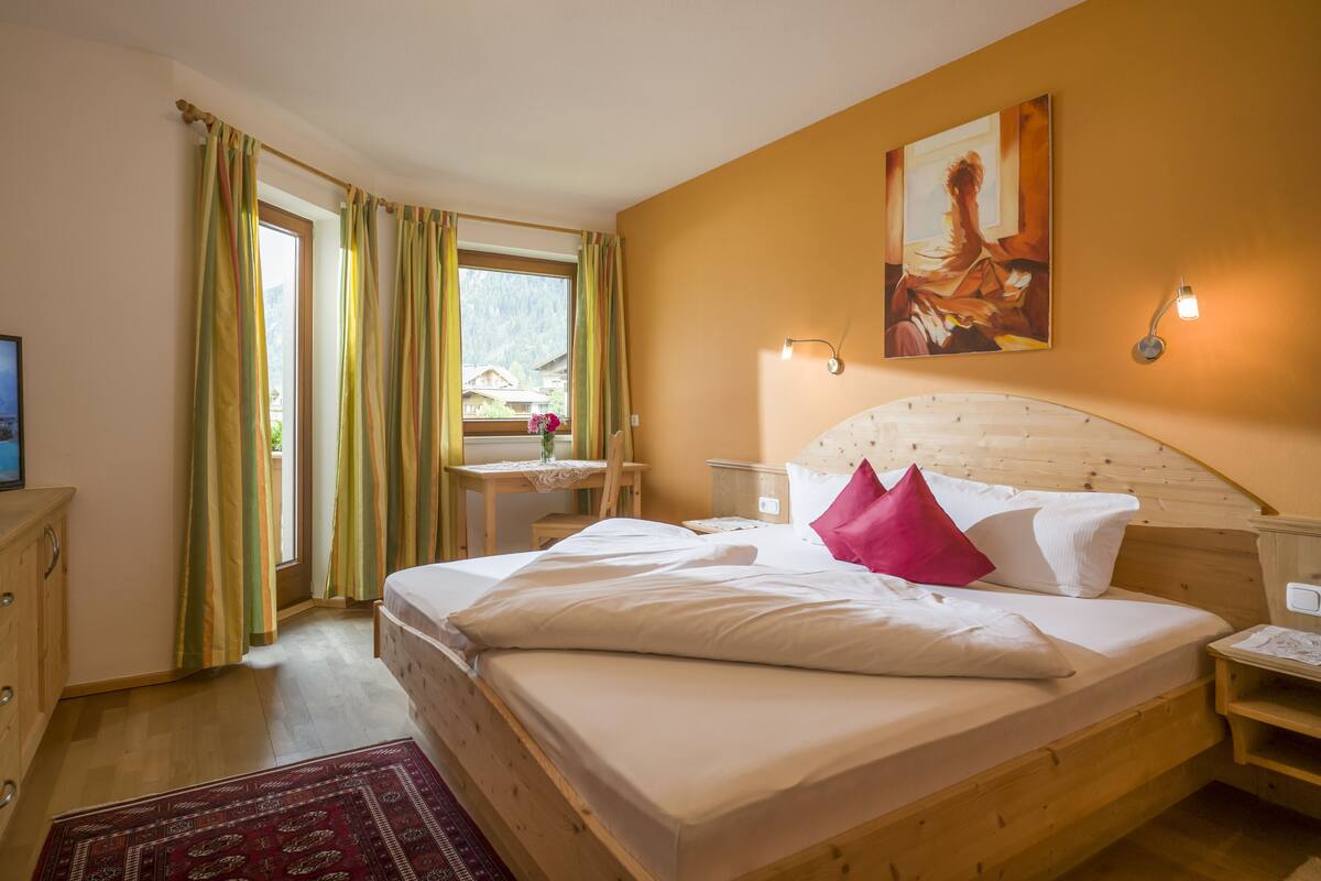 A bedroom with a bed, a desk, a chair, a TV, and a window with curtains.