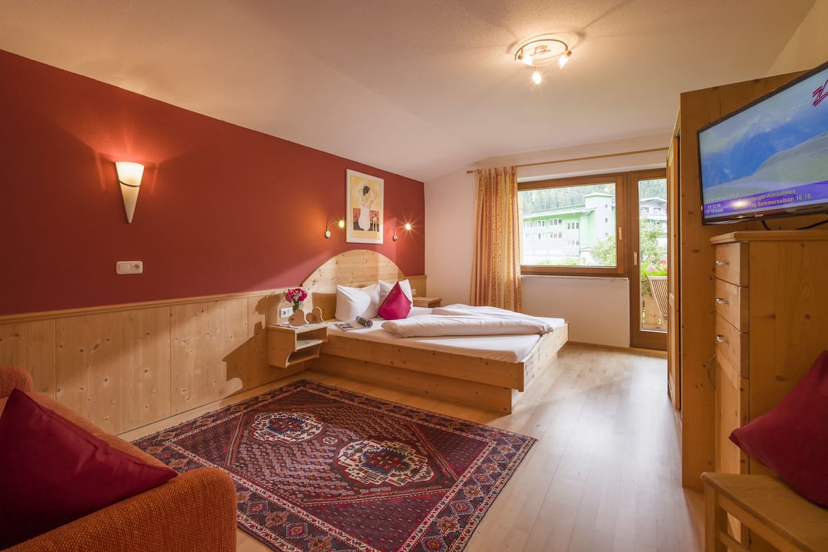 A bedroom with a wooden headboard, a bed with white linens, a red wall, a wooden dresser, a television, and a window with a view of buildings.