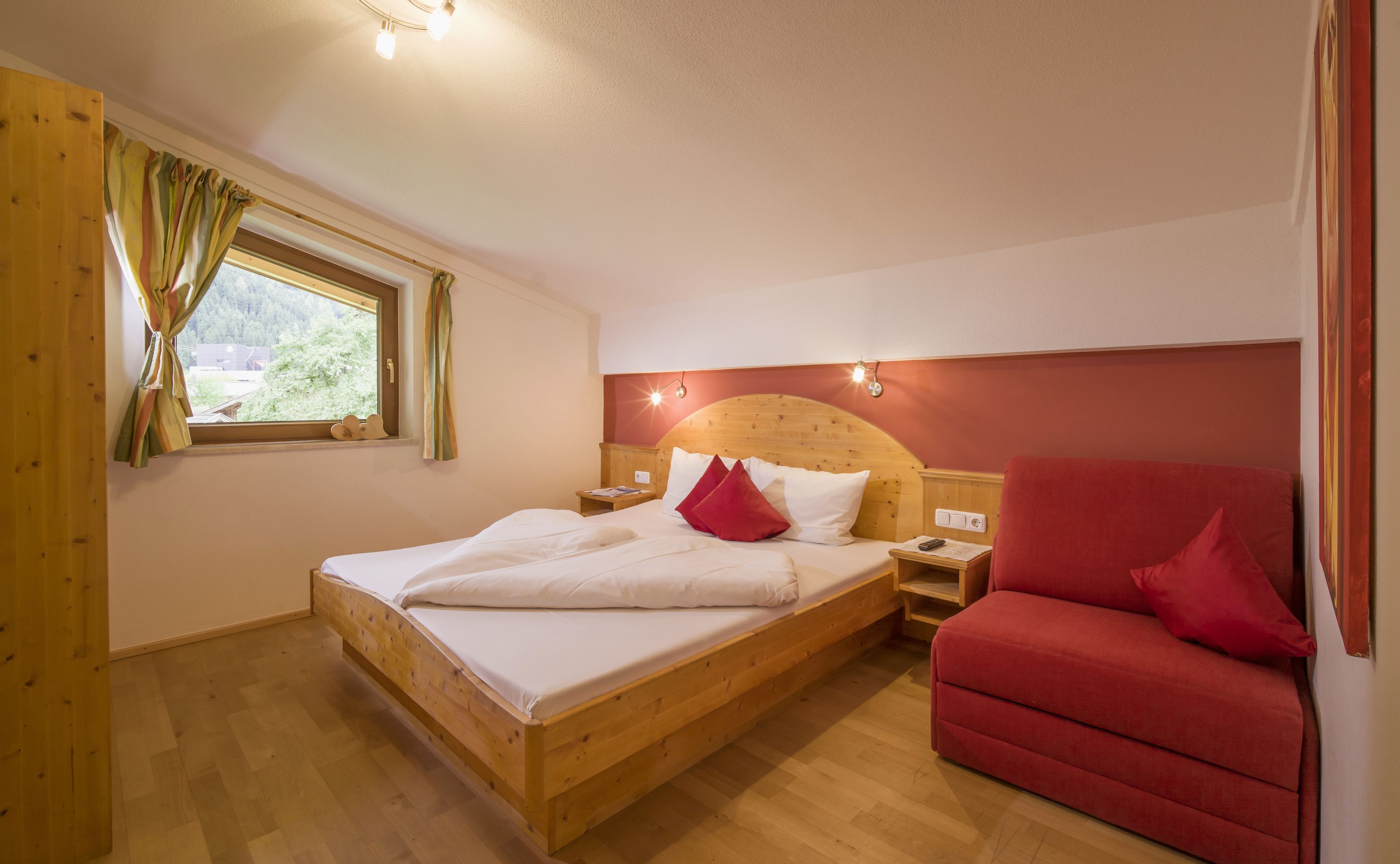 Ein Schlafzimmer mit einem hölzernen Bett, einem roten Sessel, einem Nachttisch und einem Fenster mit Vorhängen.