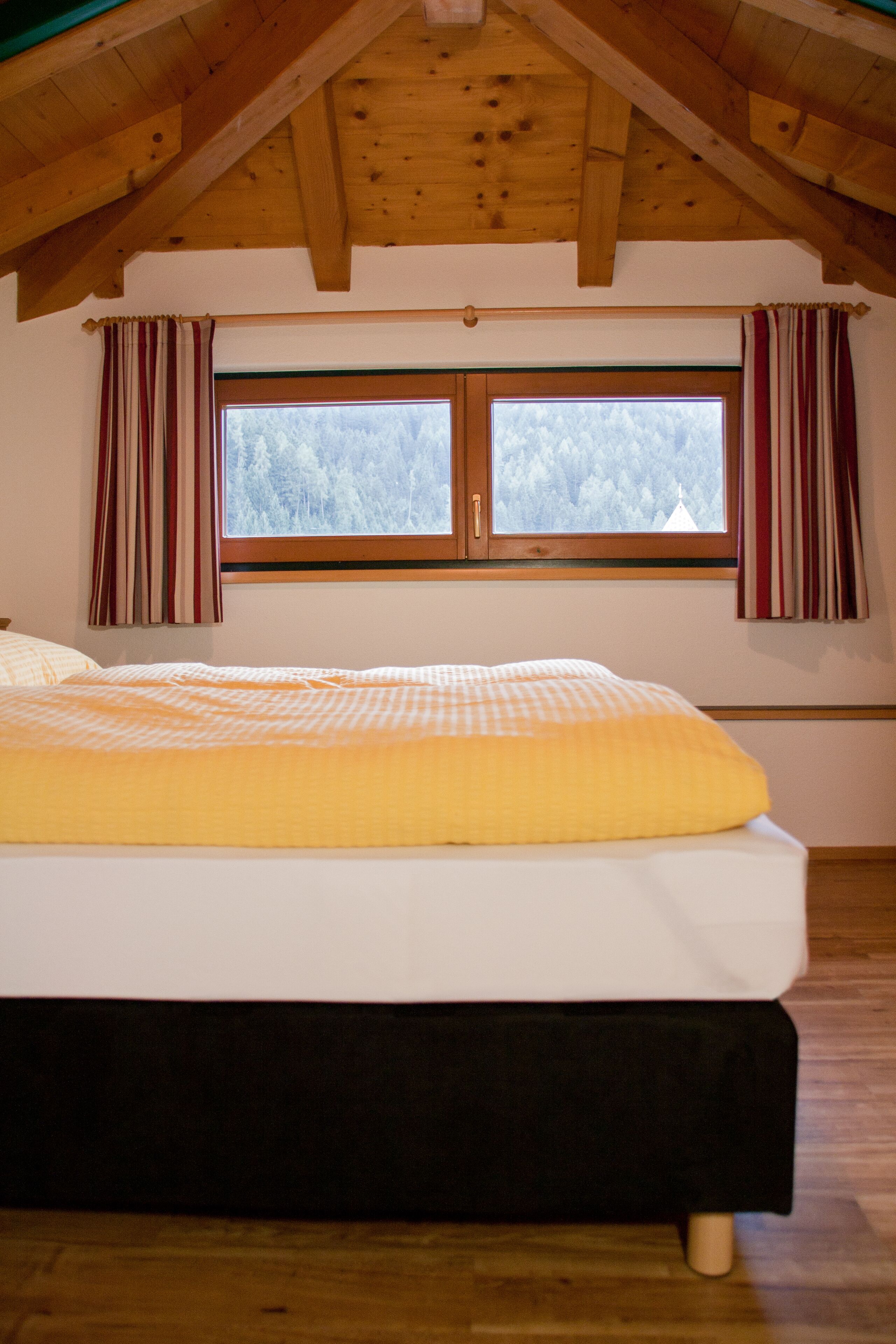 A bedroom with a large bed, wooden ceiling beams, a window with curtains, and a view of trees outside.