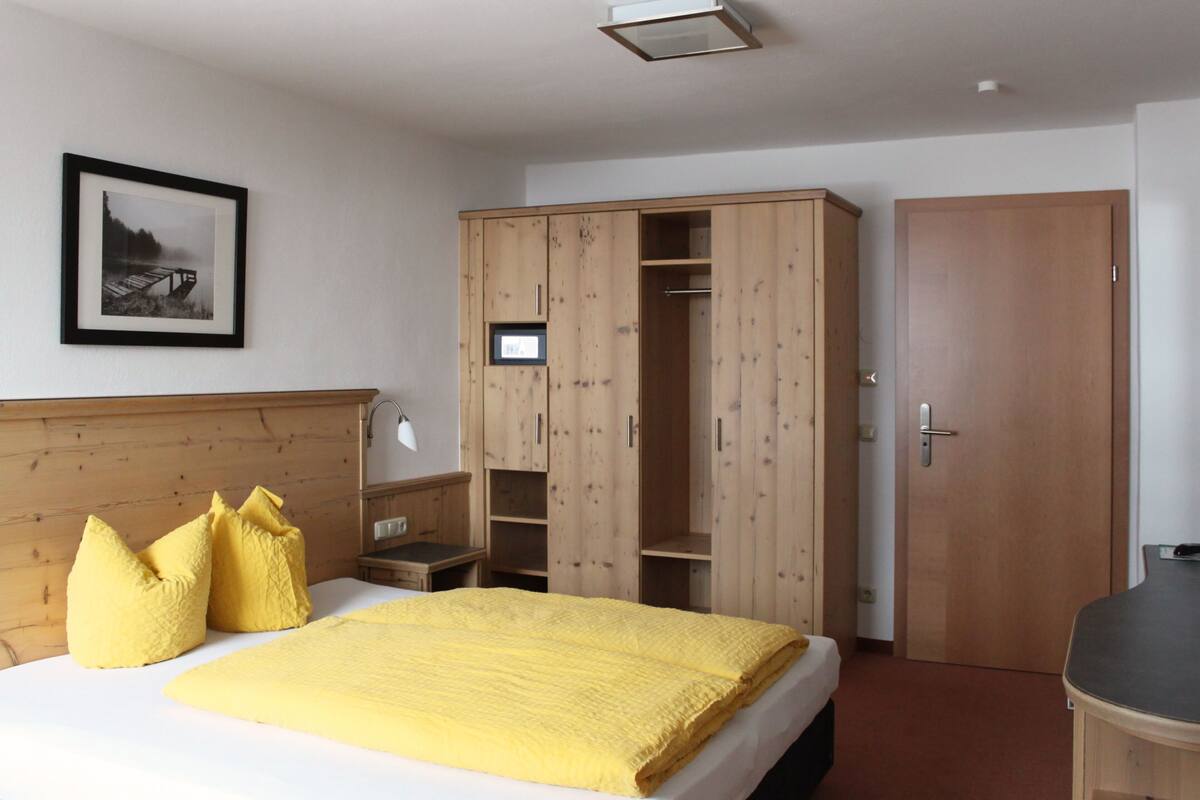 A hotel room with a bed, wooden wardrobe, bedside table, and a framed picture on the wall.