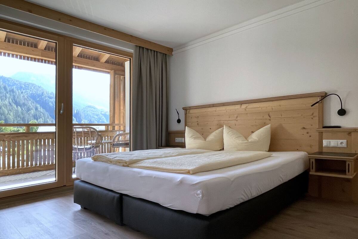 A bedroom with a wooden headboard, a large bed with white bedding, a bedside table, and a view of mountains through a sliding glass door.