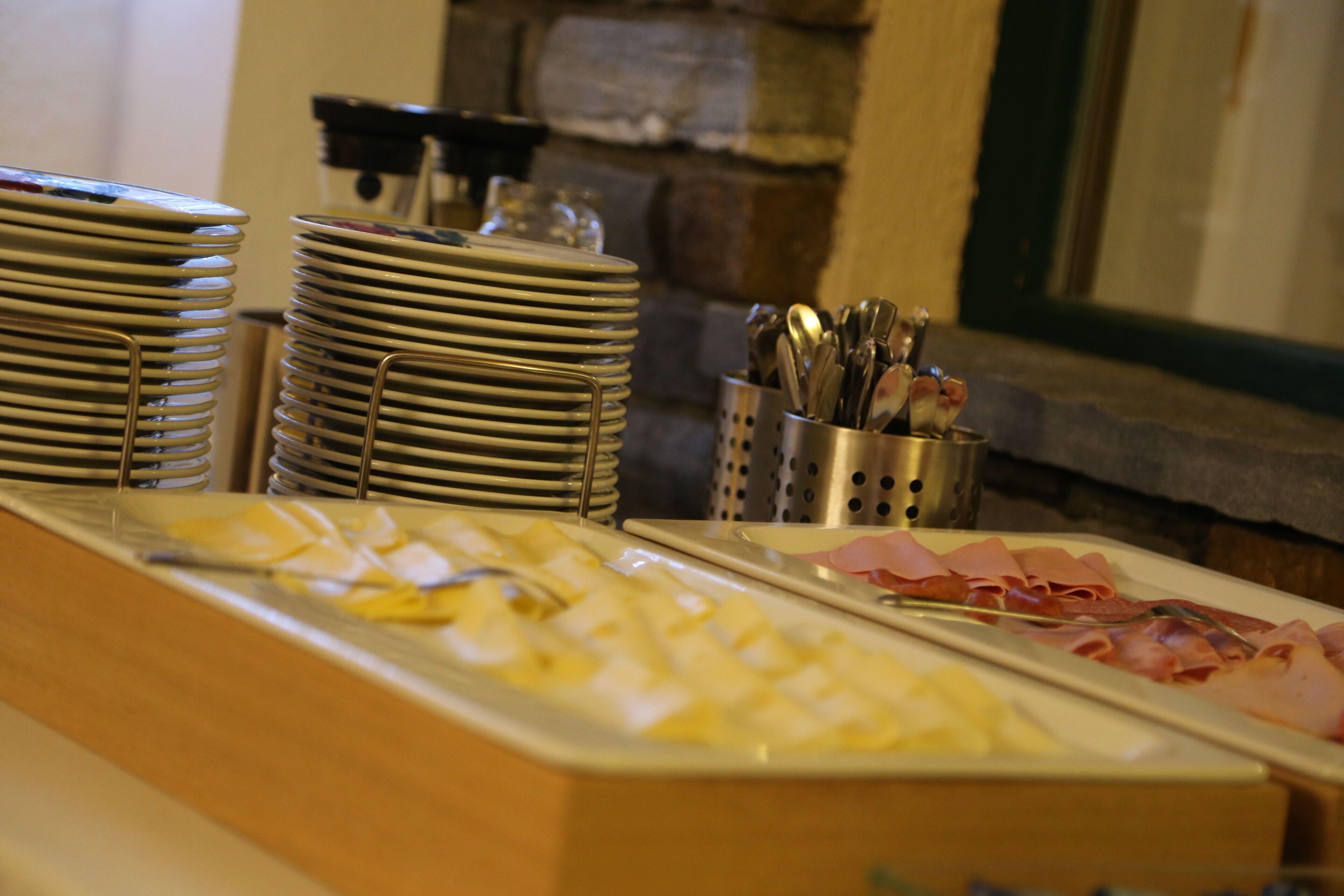 A buffet setup with stacked plates, a dish of sliced cheese, and a tray of sliced ham.