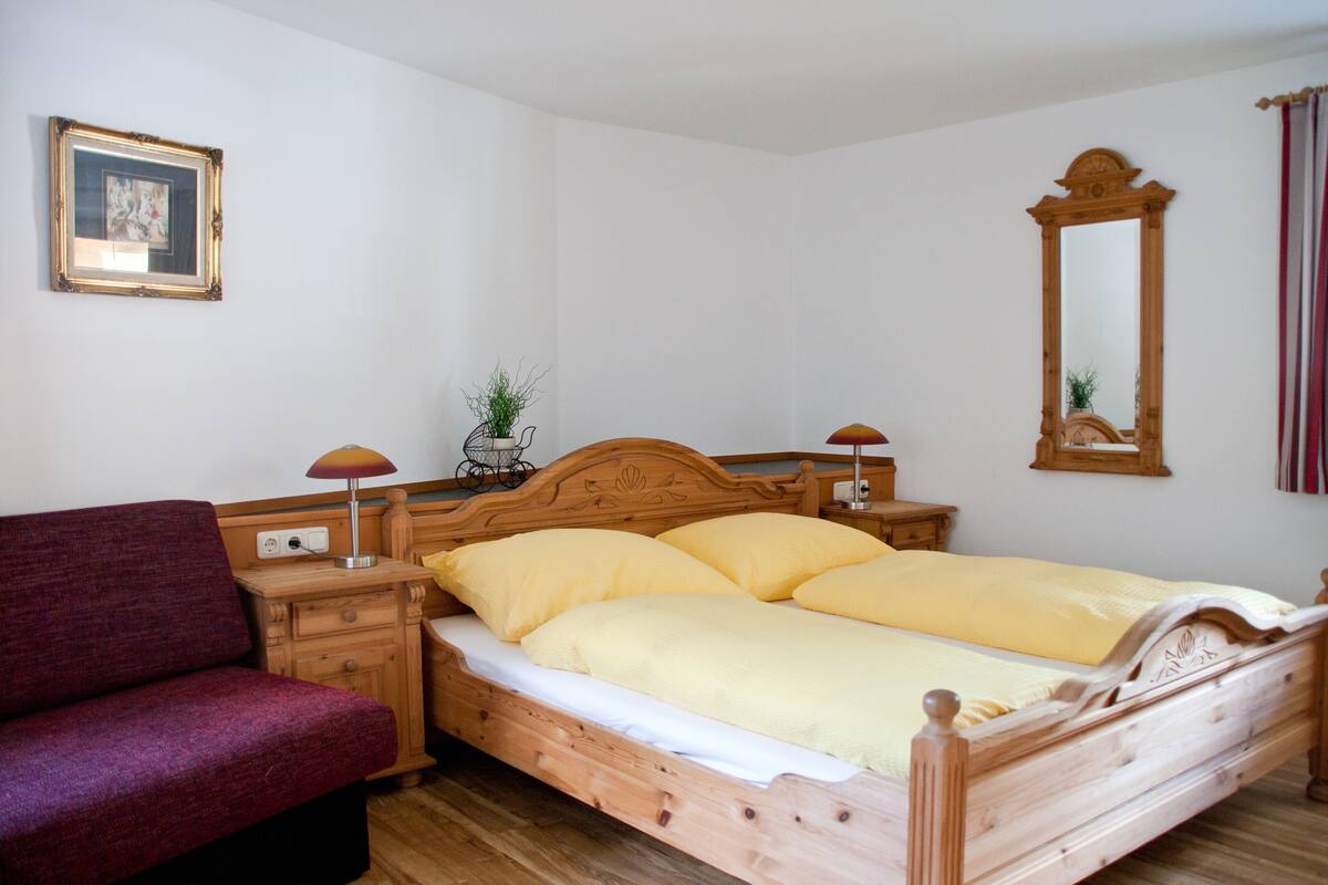 A wooden bed with yellow bedding, a wooden nightstand with a lamp, a red chair, a framed picture on the wall, and a mirror.