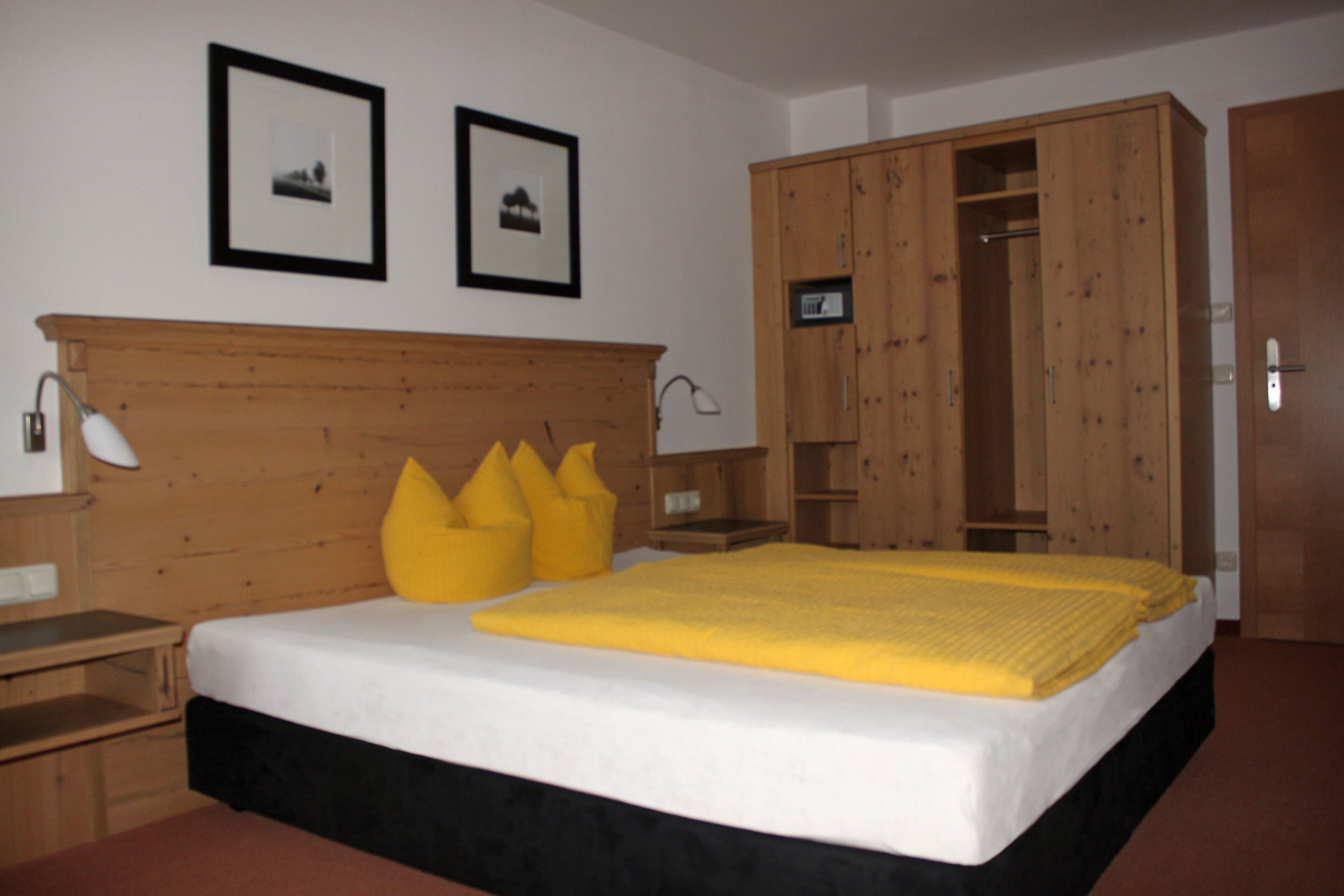 Un dormitorio con cabecera de madera, cama con manta amarilla, dos cuadros enmarcados en la pared y un armario de madera.
