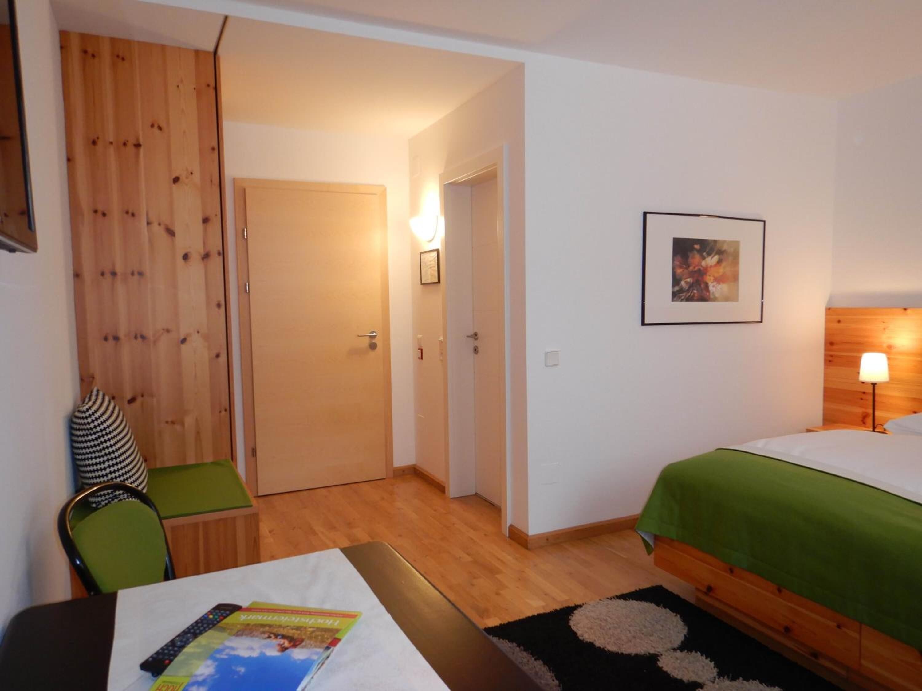 Una habitación de hotel con una cama, un escritorio con silla, un armario de madera, una puerta y una lámpara de pared.