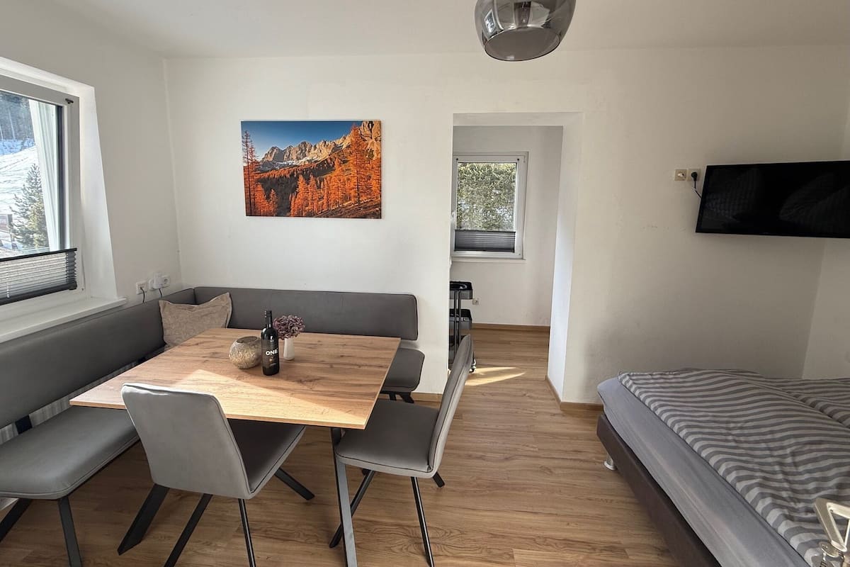 A modern, well-lit room with a wooden dining table, grey seating, a mounted TV, and a painting of a mountainous landscape.