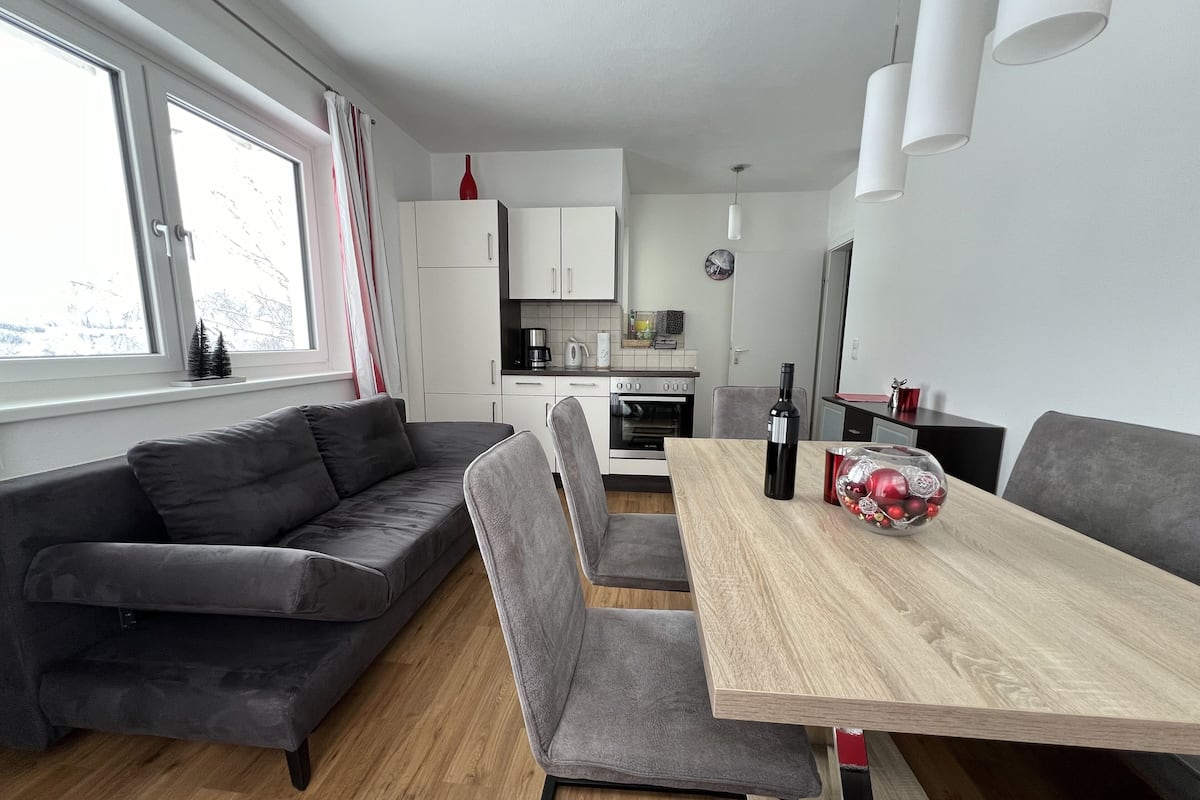 A modern kitchen with white cabinets, a wooden dining table, and a grey sofa.