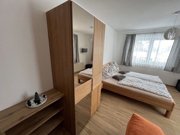 Un dormitorio moderno con cama de madera, armario empotrado, una pequeña estantería con objetos decorativos y un amplio ventanal con cortinas.