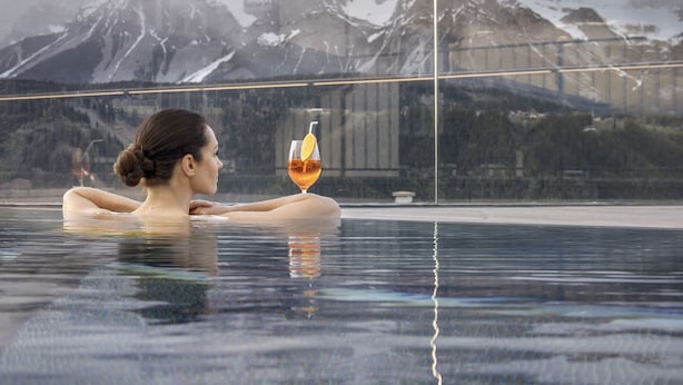 Una persona relajándose en una piscina infinita de fondo de cristal, con vista a la montaña y una pared de vidrio que muestra la palabra "ERLEBNISWELT".